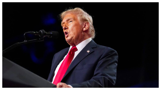 Donald Trump speaking at a podium, wearing a dark suit and red tie.